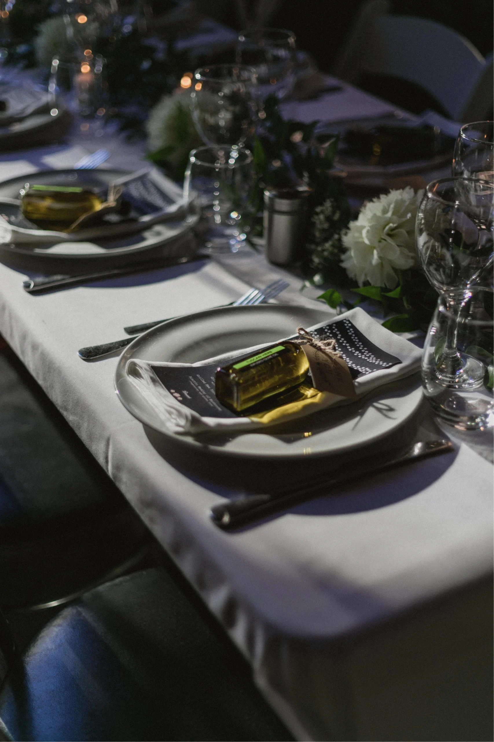 Wedding place setting with olive oil favour bottle, menu card, and floral greenery centrepiece